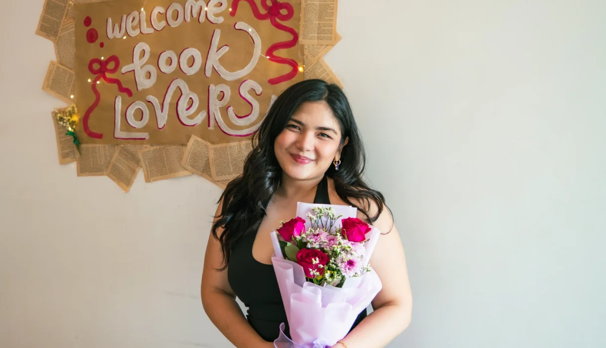 Nadz smiling and holding a bouquet of flowers in front of a welcome book lovers poster